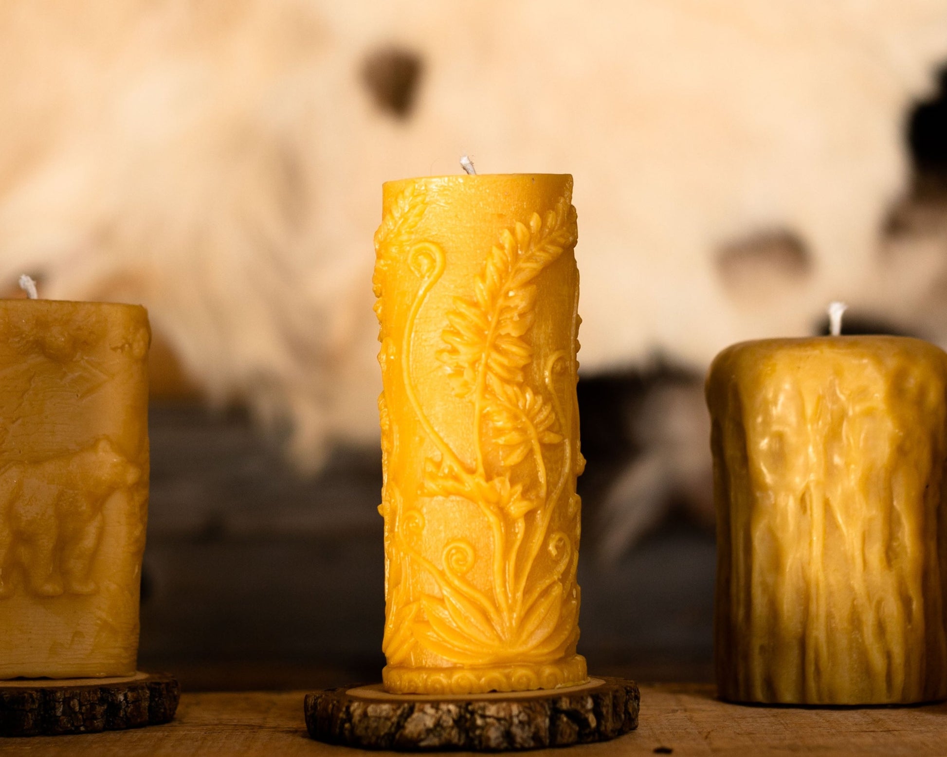 Three pillar beeswax candles with a mystic fern design, placed on wooden coasters, with a blurred animal hide in the background.